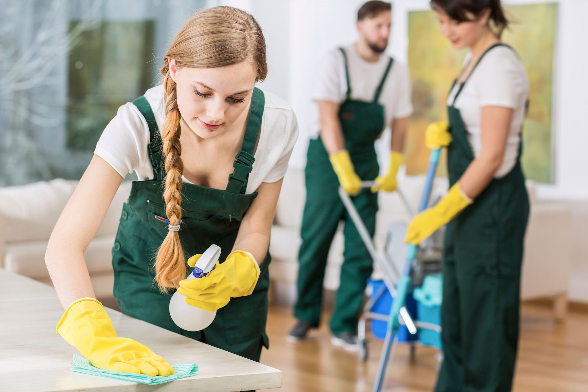 A young woman in a blue jumpsuit and green gloves is vacuuming a floor in a modern indoor space. Two other women are visible in the background, engaged in cleaning tasks. There are plants and cleaning supplies around.